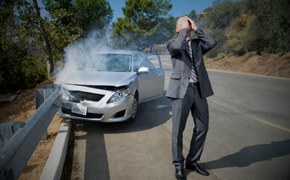 Man in suit covering his face standing near a damaged silver car crashed into a guardrail with smoke.